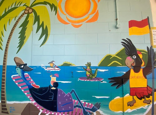 street art image of a black cockatoo standing on a surfboard in the yellow/red lifesaver uniform and cassowary on a recliner on a tropical beach and crocodile standup paddleboarding in the ocean